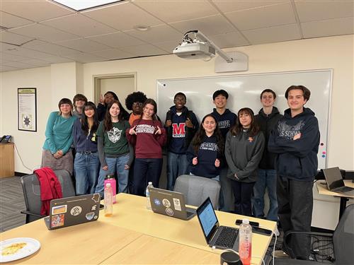 Group of diverse students standing in front of a white board and projector smiling at the camera.